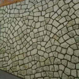 Ściana z nieregularnych, jasnych kamieni, ułożonych w łukowaty wzór, z małym oknem po lewej stronie i fragmentem drewnianej konstrukcji u góry. Widoczny fragment chodnika i nieba.