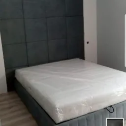 Szare tapicerowane łóżko z wysokim zagłówkiem podzielonym na kwadraty, materac owinięty folią ochronną, widok z boku w jasnym pomieszczeniu.