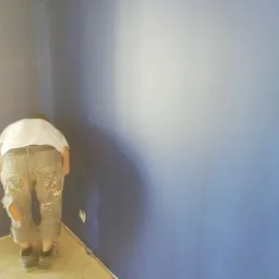 Mężczyzna w roboczych spodniach, pochylony, maluje niebieską ścianę w rogu pomieszczenia, widoczne zabezpieczone podłogi i gniazdko elektryczne.