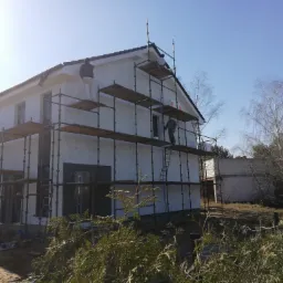 Dwukondygnacyjny dom w trakcie prac wykończeniowych elewacji, otoczony rusztowaniem, z pracownikami na różnych poziomach, w słoneczny dzień.