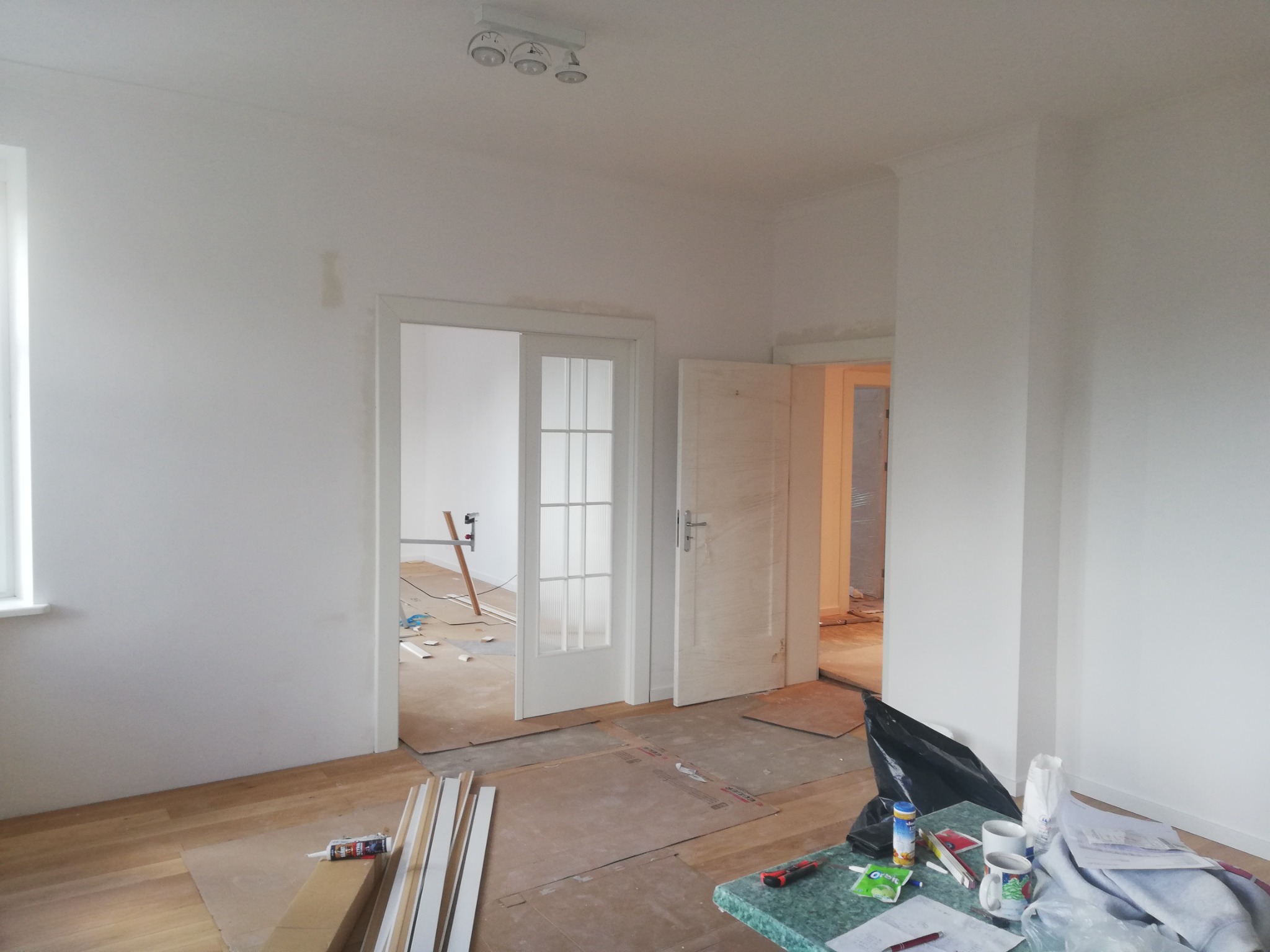 Pokój w trakcie remontu: widoczne białe ściany, podłoga częściowo zabezpieczona kartonem, listwy przypodłogowe, otwarte drzwi wewnętrzne z szybami, narzędzia na stole, oraz ślady po naprawach...