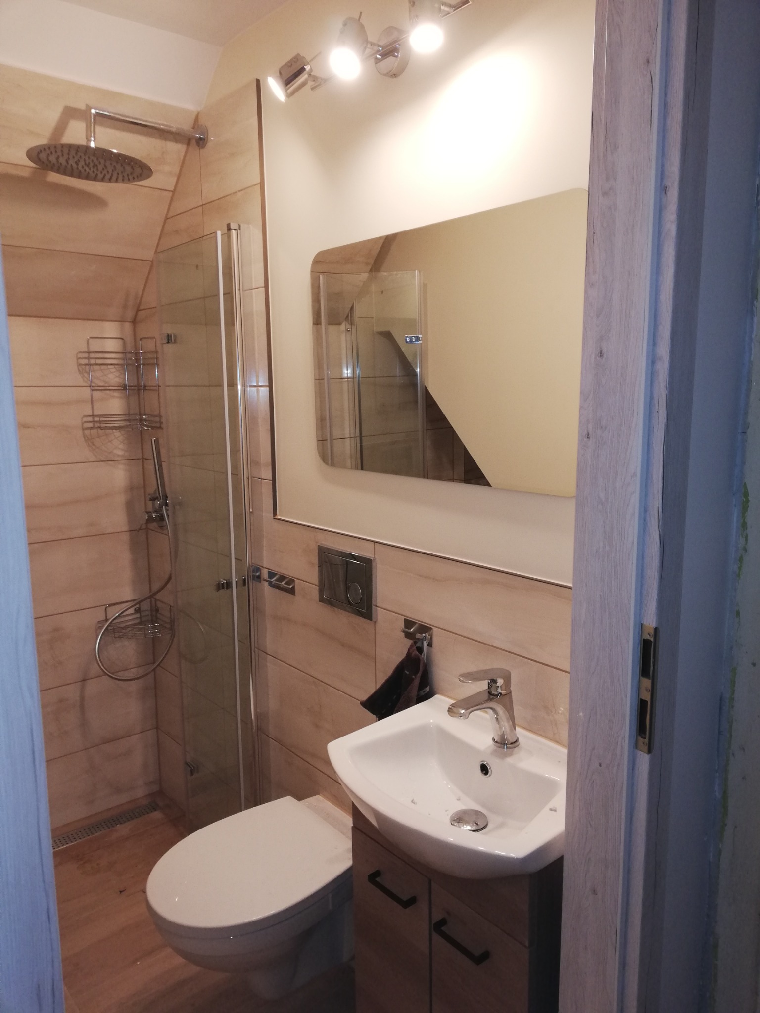 Nowoczesna łazienka z kabiną prysznicową typu walk-in, umywalką nablatową na drewnianej szafce, dużym lustrem i oświetleniem punktowym, widziana zza częściowo otwartych drzwi.