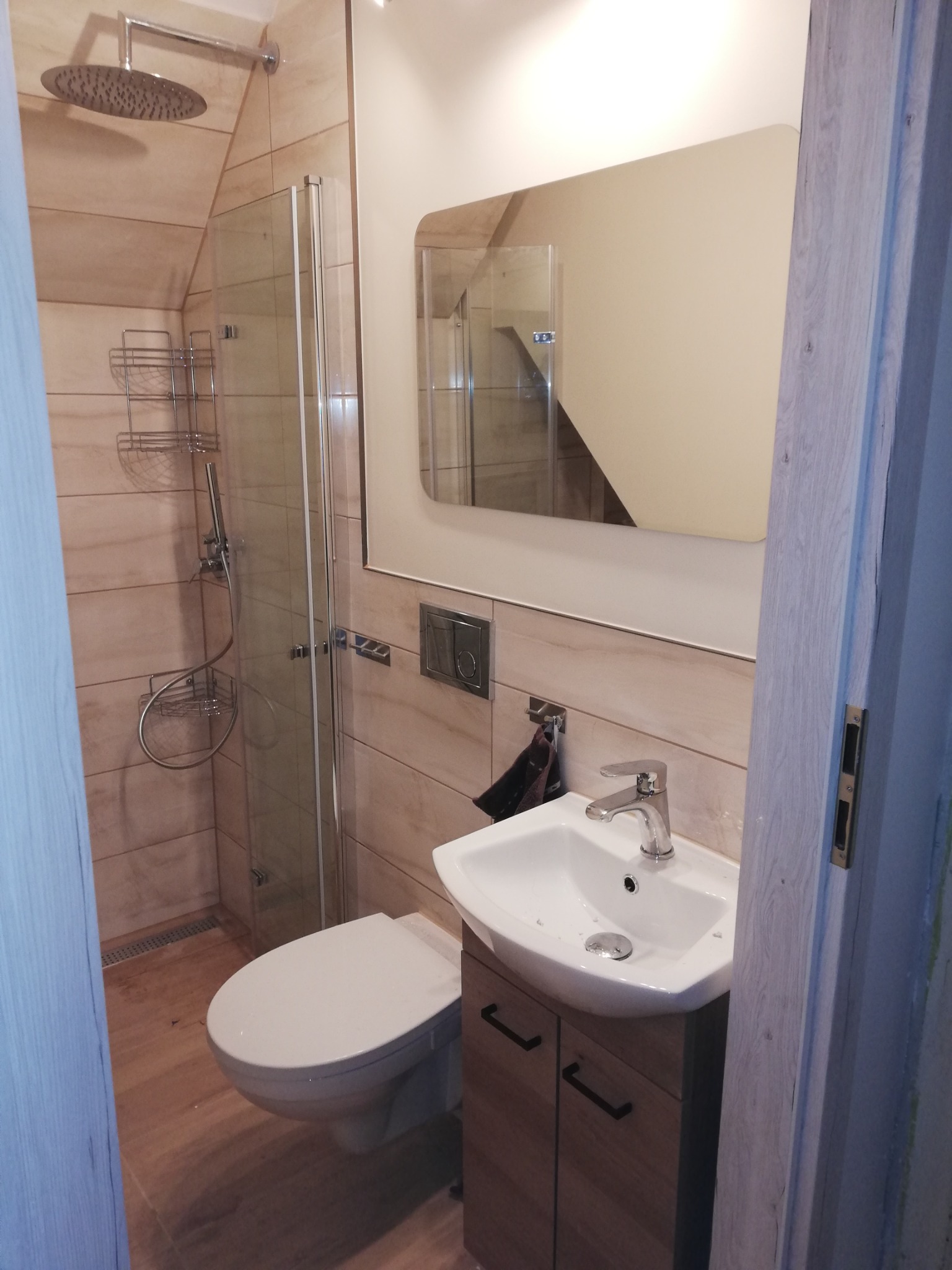 Kompletnie wykończona łazienka na poddaszu z beżowymi płytkami imitującymi drewno, kabiną prysznicową ze szklanymi drzwiami, umywalką z szafką w kolorze drewna i wiszącą toaletą.