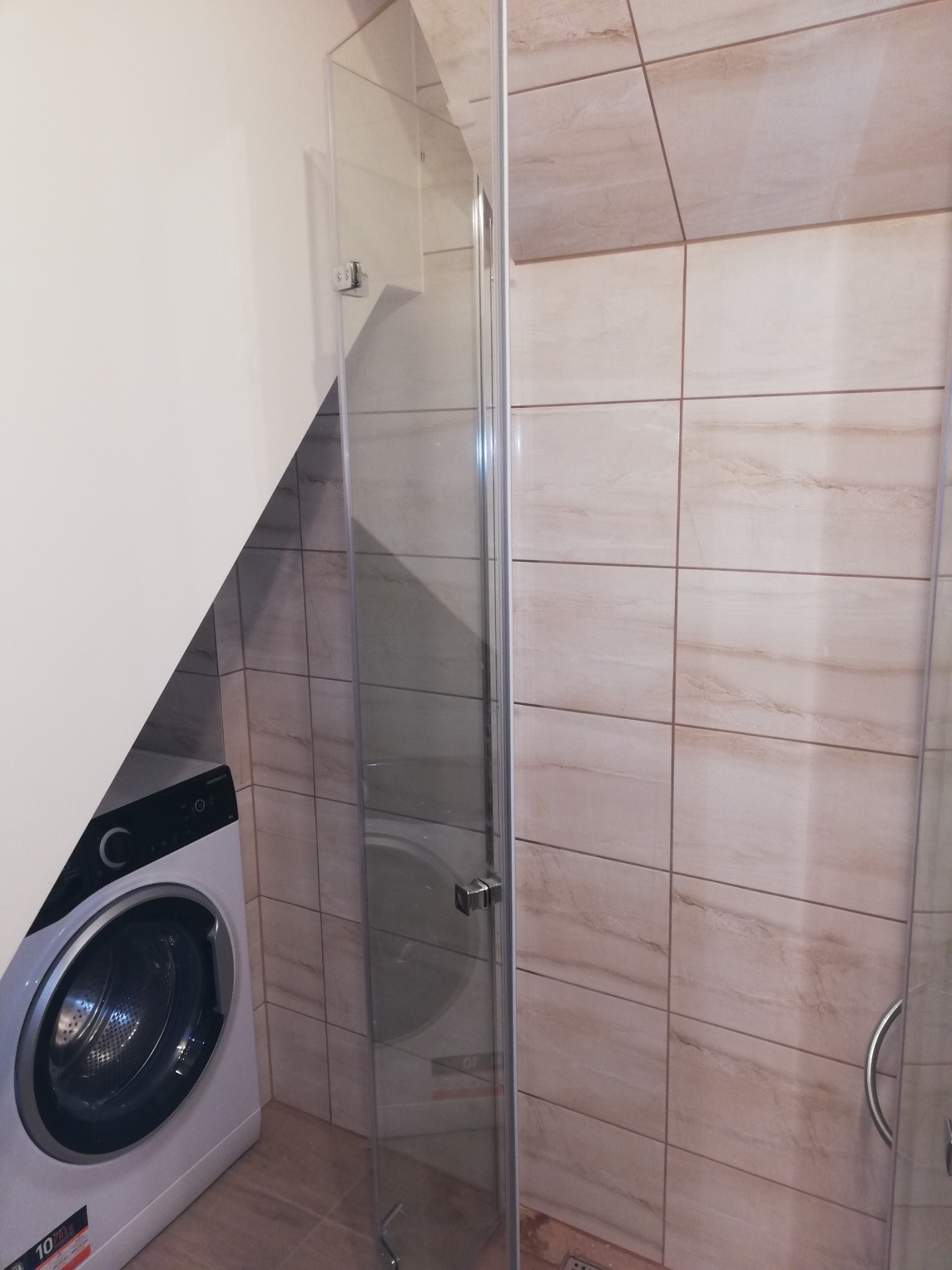 Łazienka z kabiną prysznicową ze szkła hartowanego, beżowe płytki imitujące marmur na ścianach, pralka pod skosem dachu.