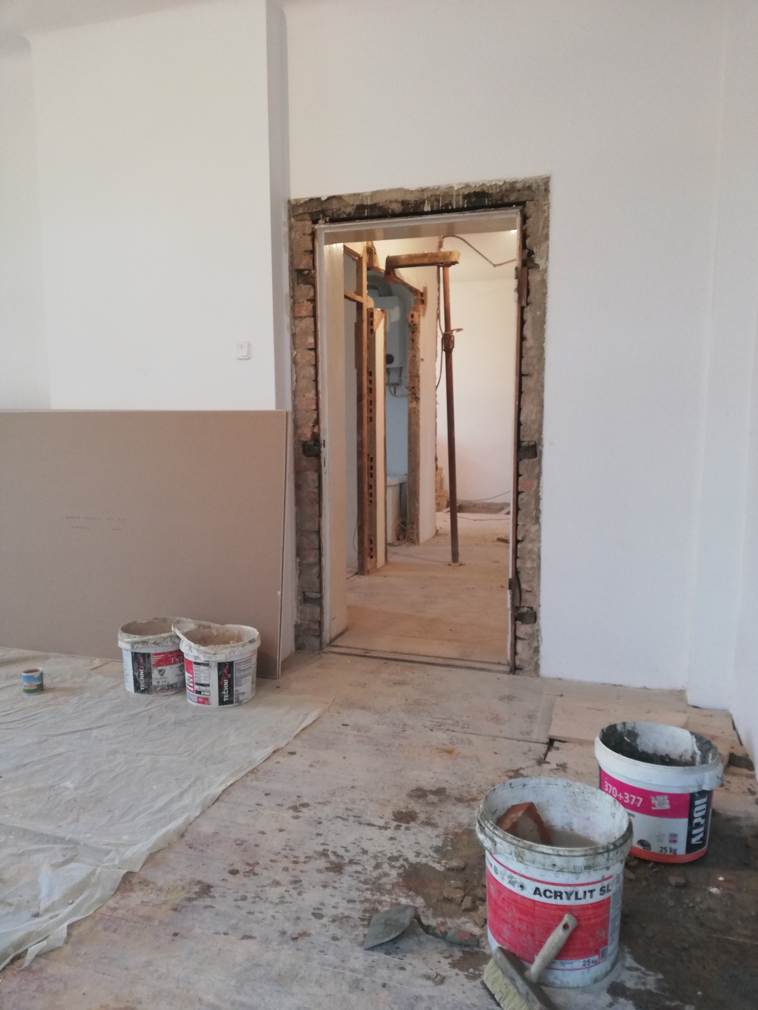 Wnętrze w trakcie remontu: widoczne płyty gipsowe, otwarte puszki z farbą, narzędzia malarskie i surowa ościeżnica drzwiowa z odsłoniętą cegłą, w tle widoczne rusztowania i podtrzymujące stemple.