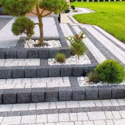 Schody i ścieżka z kostki brukowej w dwóch odcieniach szarości, z elementami dekoracyjnymi w postaci białych kamieni i roślin, prowadzące do domu z zadbanym trawnikiem w tle.