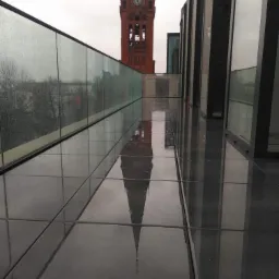 Błyszcząca, mokra posadzka balkonu z widokiem na wieżę zegarową odbijającą się w jej powierzchni, przeszklona balustrada z kroplami deszczu.