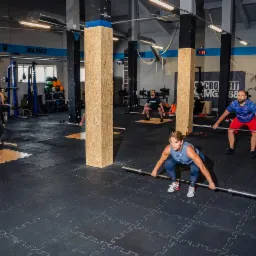 Trening siłowy w siłowni: grupa osób wykonuje przysiady ze sztangą, widoczne słupy obłożone płytami OSB, gumowa podłoga w kształcie puzzli.
