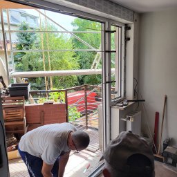 POL-DRZWI - Montaż okna PCV w Bystrem: Pracownik z wiertarką mocuje ramę okienną. Widoczne rusztowania i zieleń za oknem. Wnętrze w trakcie prac wykończeniowych.