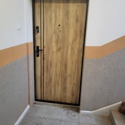 POL-DRZWI - Stylowe drzwi wejściowe z drewnianym wzorem i czarną ramą, zamontowane w klatce schodowej z szaro-pomarańczowymi ścianami. Widoczne detale zamka i wizjera.