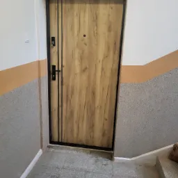 Stylowe drzwi wejściowe z drewnianym wzorem i czarną ramą, zamontowane w klatce schodowej z szaro-pomarańczowymi ścianami. Widoczne detale zamka i wizjera.