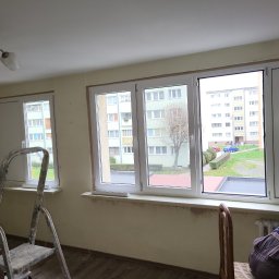 POL-DRZWI - Wnętrze z trzema oknami PCV w trakcie montażu. Widoczna drabina, odsłonięte ściany i widok na blok z zewnątrz. Częściowo widoczny pracownik w kombinezonie.