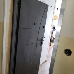 POL-DRZWI - Ciemne, nowoczesne drzwi z metalowymi akcentami, zamontowane w ościeżnicy. Widoczny fragment wnętrza z białymi szafkami i osobą w tle. Ściana z otworem na instalację.
