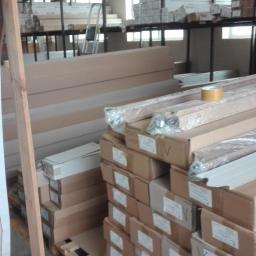 POL-DRZWI - Magazyn pełen zapakowanych elementów stolarki budowlanej, widoczne kartony na paletach, półki z towarem i drabina w tle.