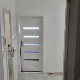 Białe drzwi wewnętrzne z poziomymi przeszkleniami w otwartym mieszkaniu, widok z perspektywy wejścia do pomieszczenia, z fragmentem otwartych drzwi po lewej stronie i stosem paneli podłogowych przy...