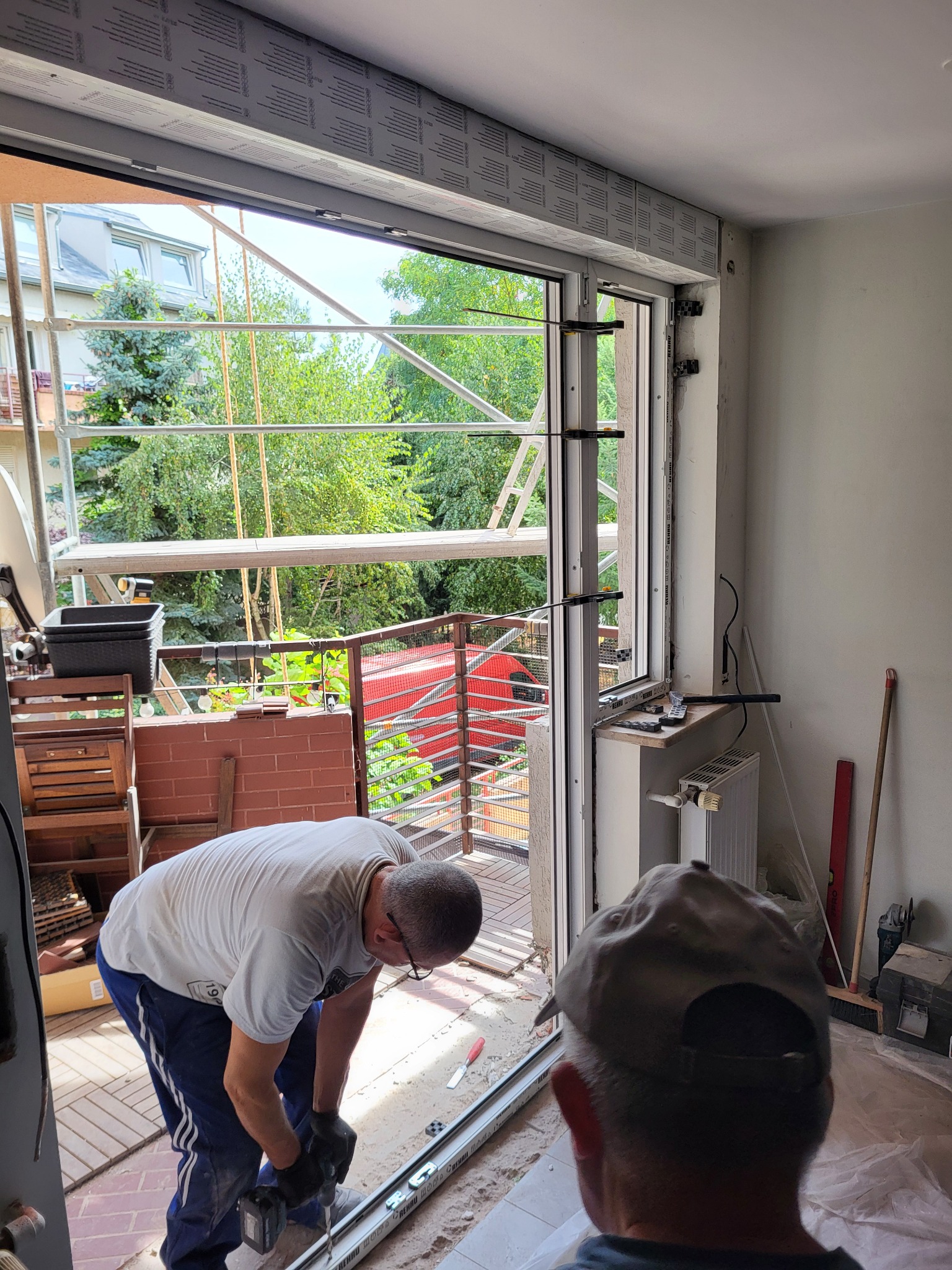 Montaż okna PCV w Bystrem: Pracownik z wiertarką mocuje ramę okienną. Widoczne rusztowania i zieleń za oknem. Wnętrze w trakcie prac wykończeniowych.