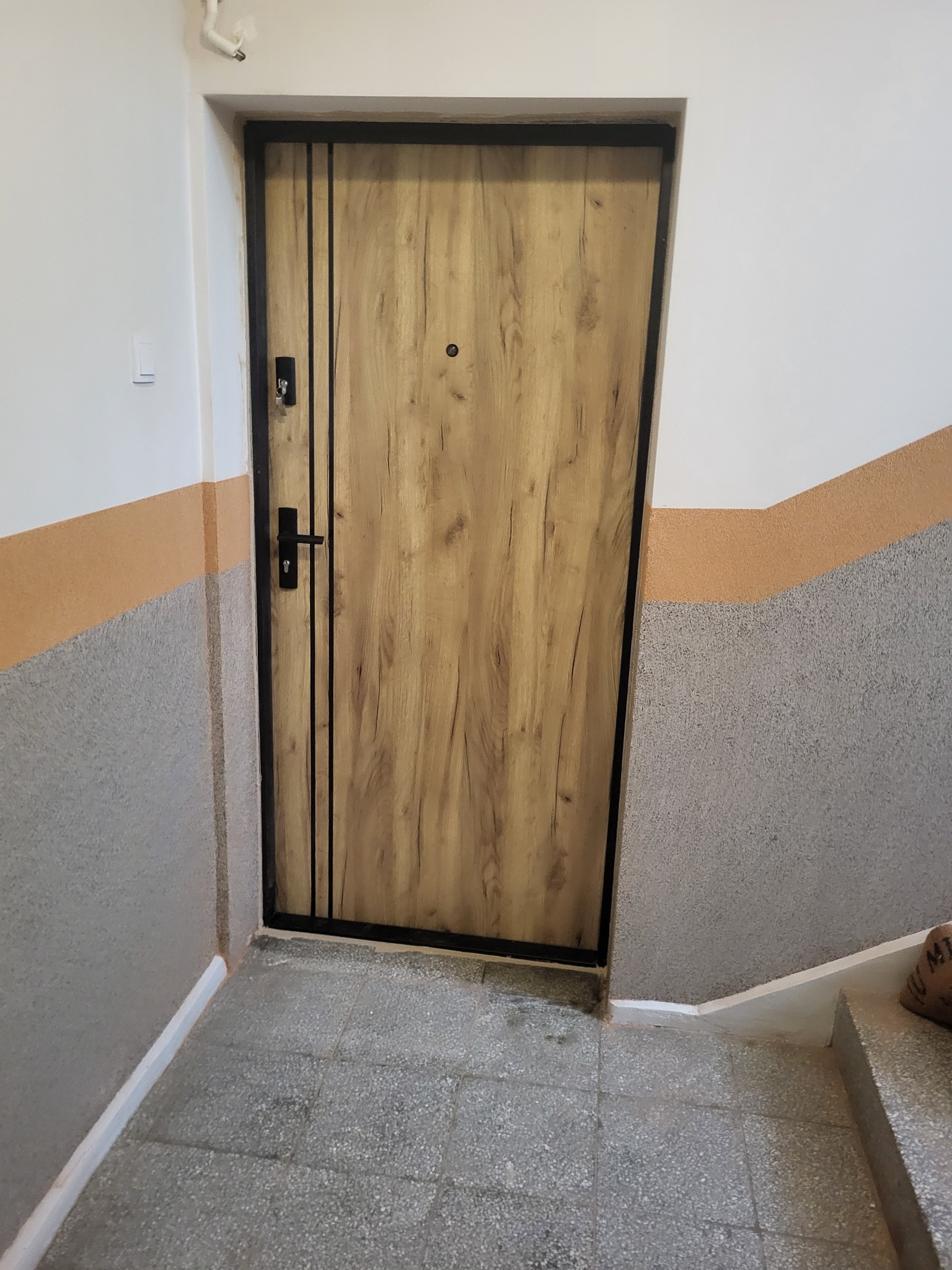 Stylowe drzwi wejściowe z drewnianym wzorem i czarną ramą, zamontowane w klatce schodowej z szaro-pomarańczowymi ścianami. Widoczne detale zamka i wizjera.