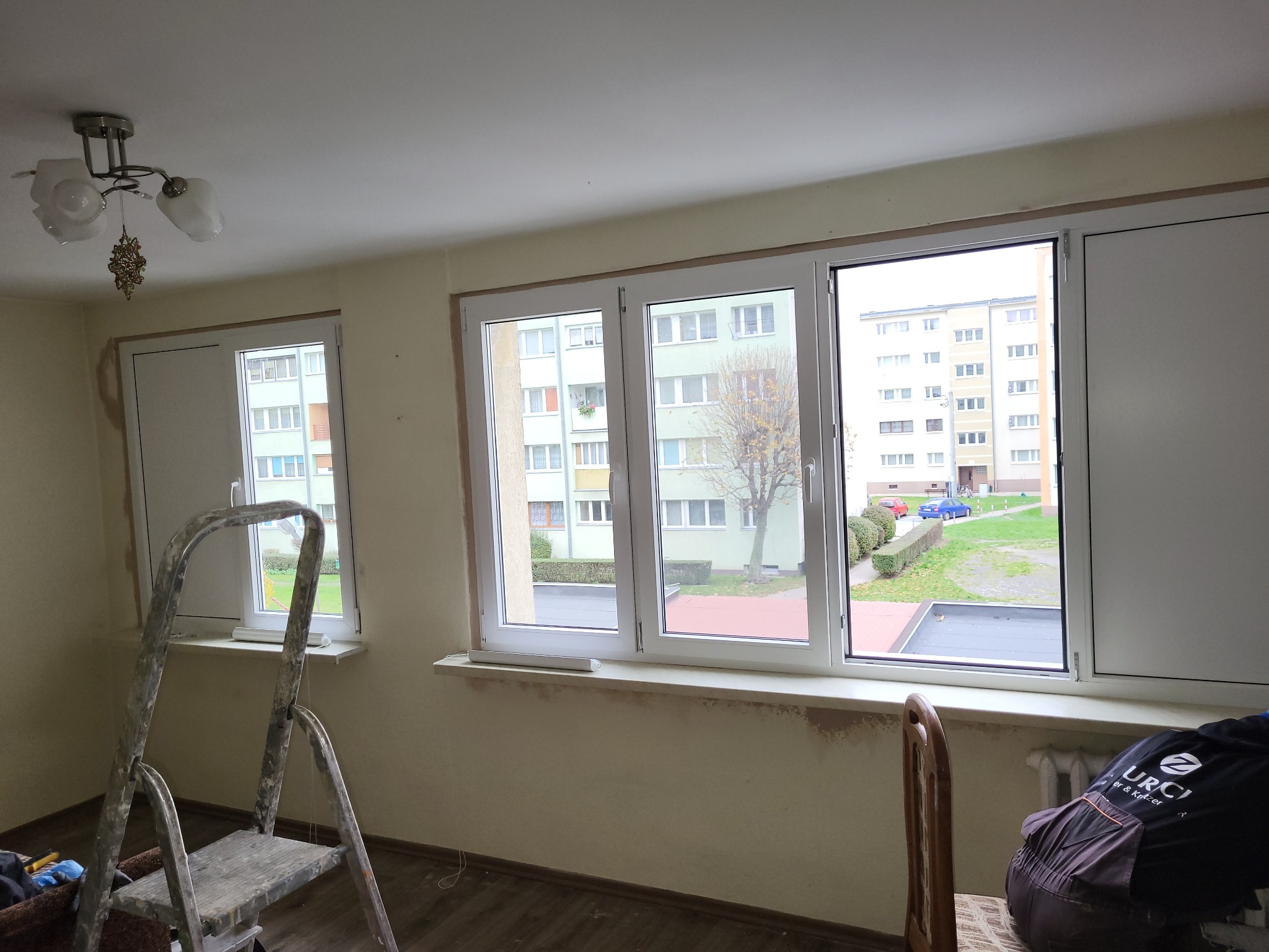Wnętrze z trzema oknami PCV w trakcie montażu. Widoczna drabina, odsłonięte ściany i widok na blok z zewnątrz. Częściowo widoczny pracownik w kombinezonie.