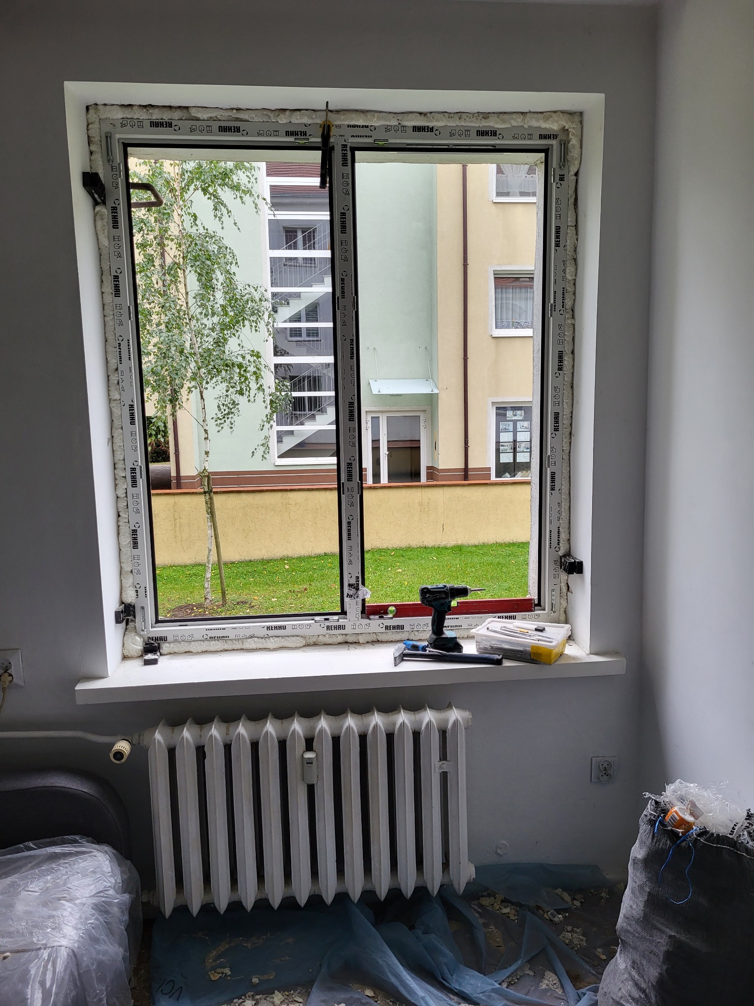Montaż okna PCV Rehau z widokiem na zieleń. Pianka montażowa uszczelnia ramę. Na parapecie narzędzia: wkrętarka, młotek i nożyk. Grzejnik pod oknem. Bałagan po montażu.