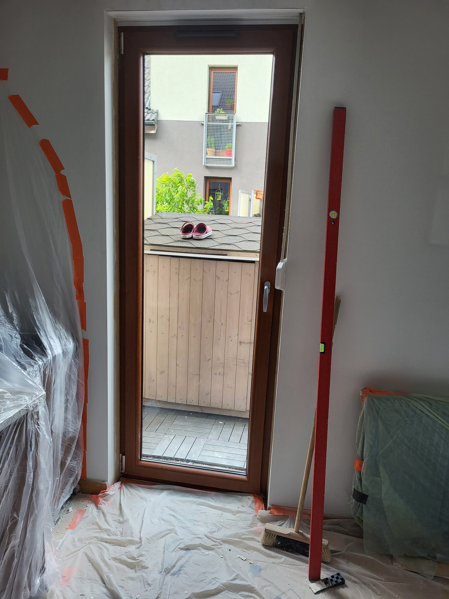 Montaż brązowych drzwi balkonowych. Poziomica oparta o ścianę. Podłoga zabezpieczona folią malarską. Na dachu sąsiedniego balkonu leżą różowe klapki.