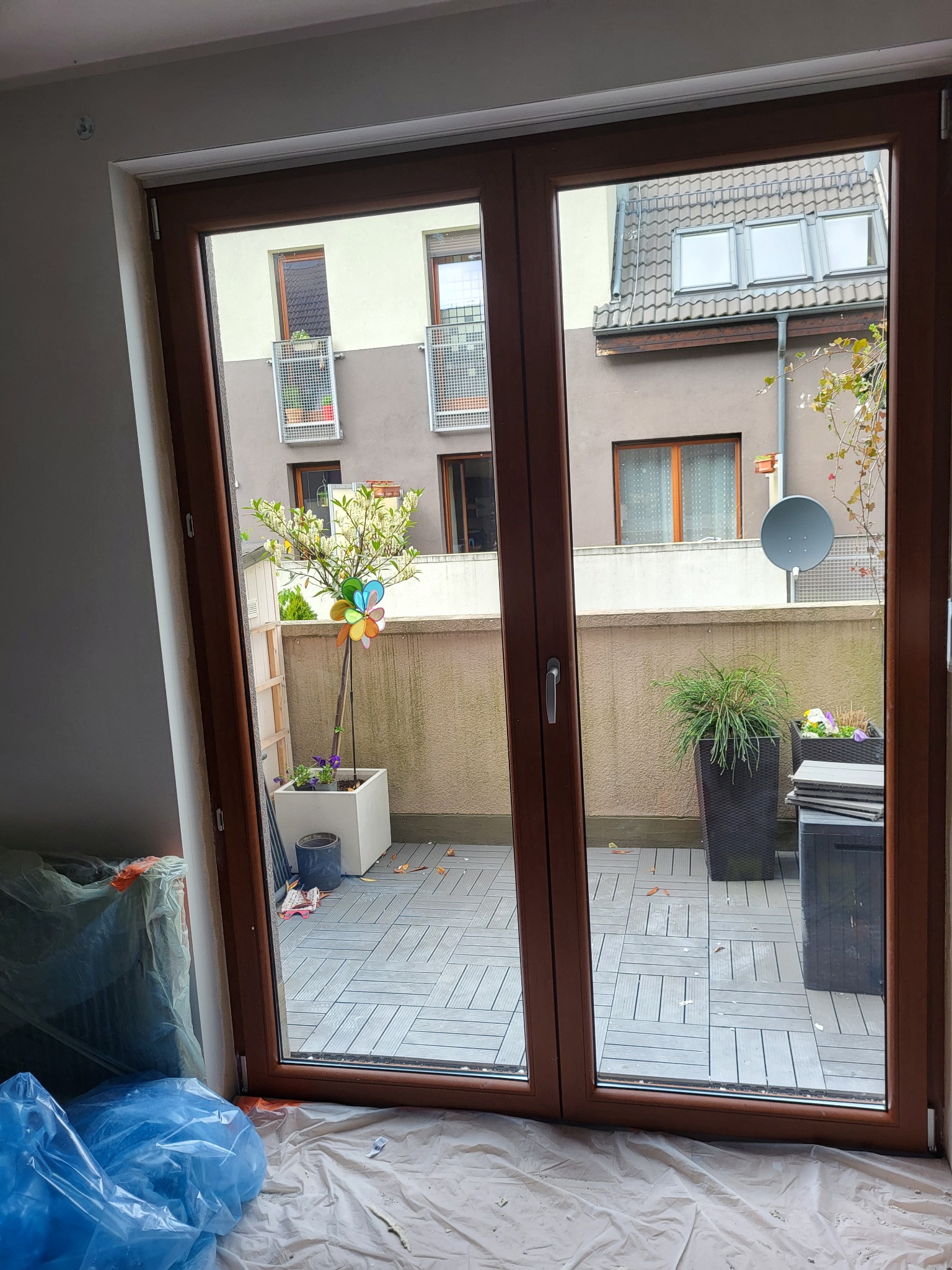 Brązowe dwuskrzydłowe okno balkonowe z widokiem na taras i sąsiedni budynek. Okno zabezpieczone folią malarską, widoczne prace wykończeniowe.