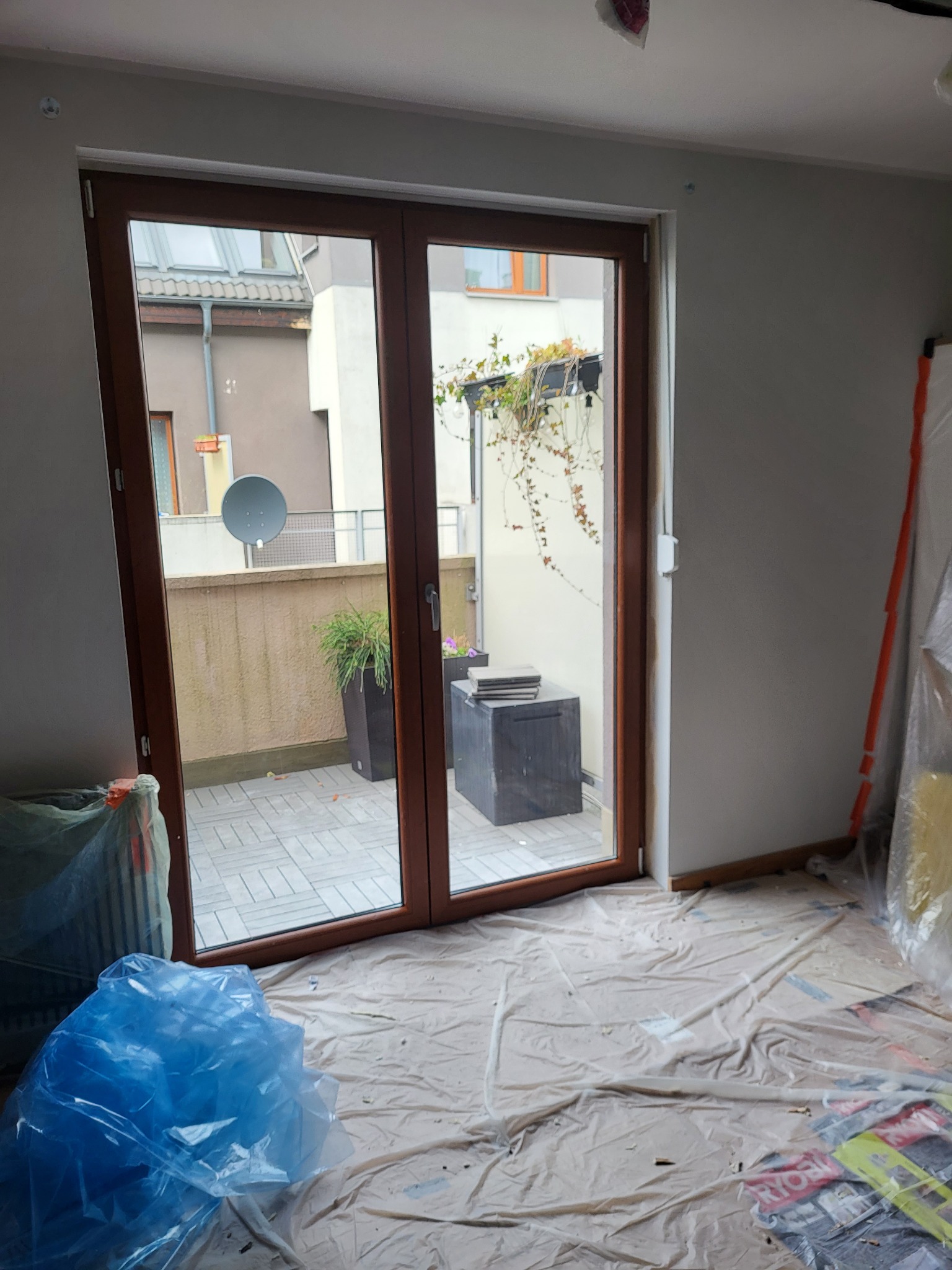 Brązowe, dwuskrzydłowe drzwi balkonowe z widokiem na taras. Podłoga zabezpieczona folią malarską, ściany szare. Widoczne rośliny w donicach i fragment zabudowy.