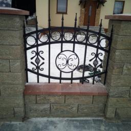 Dark Metal - Fragment ogrodzenia z czarnym, metalowym przęsłem o dekoracyjnym wzorze, osadzonego między kamiennymi słupkami z ceglaną wstawką, na tle ośnieżonego trawnika i domu.