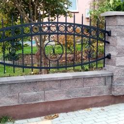 Dark Metal - Fragment ogrodzenia z czarną metalową balustradą o dekoracyjnych elementach, osadzoną na murku z szarej kostki i słupkach z tego samego materiału, widok z zewnątrz na tle zieleni ogrodu.