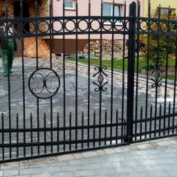 Dark Metal - Czarna, dwuskrzydłowa brama z dekoracyjnymi elementami, widoczna z bliska na tle kostki brukowej i budynku mieszkalnego z ułożonym drewnem za nią, podczas prac montażowych.