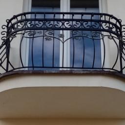 Dark Metal - Kuty balkon z dekoracyjnymi elementami roślinnymi, widok z dołu na tle okna z odbijającym się niebem.