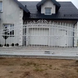 Biała, ozdobna brama wjazdowa z metalu, z kwiatowymi motywami, na tle domu z garażem i starannie utrzymanym trawnikiem, widziana z perspektywy ziemi.