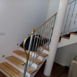 Dark Metal - Wnętrze domu z drewnianymi schodami i metalową balustradą z elementami w kolorze złotym. Na schodach widoczne zabezpieczenia budowlane.