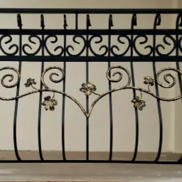 Czarna balustrada balkonowa z dekoracyjnym motywem roślinnym w kolorze złotym, widoczna na tle jasnej ściany.