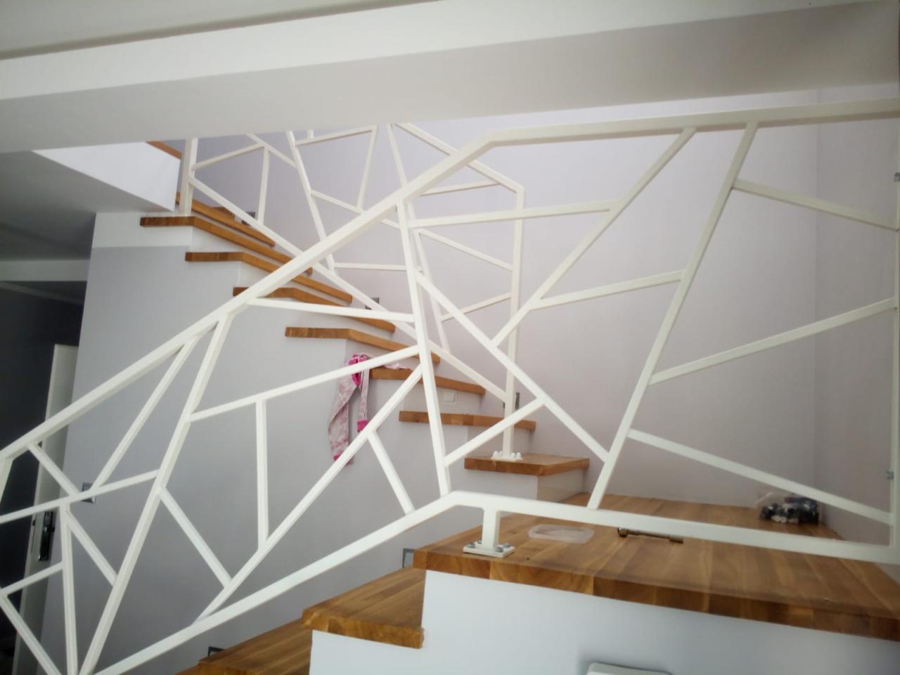 Schody z drewnianymi stopniami i białą, geometryczną konstrukcją zabezpieczającą, przypominającą nowoczesną instalację artystyczną.