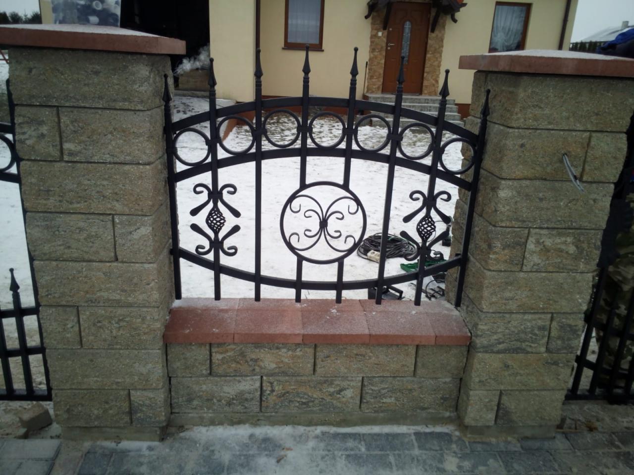 Fragment ogrodzenia z czarnym, metalowym przęsłem o dekoracyjnym wzorze, osadzonego między kamiennymi słupkami z ceglaną wstawką, na tle ośnieżonego trawnika i domu.