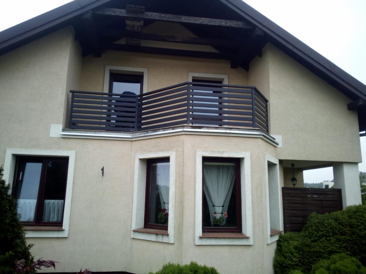 Szara, pozioma balustrada na balkonie narożnym domu jednorodzinnego z beżową elewacją i brązowymi ramami okiennymi.