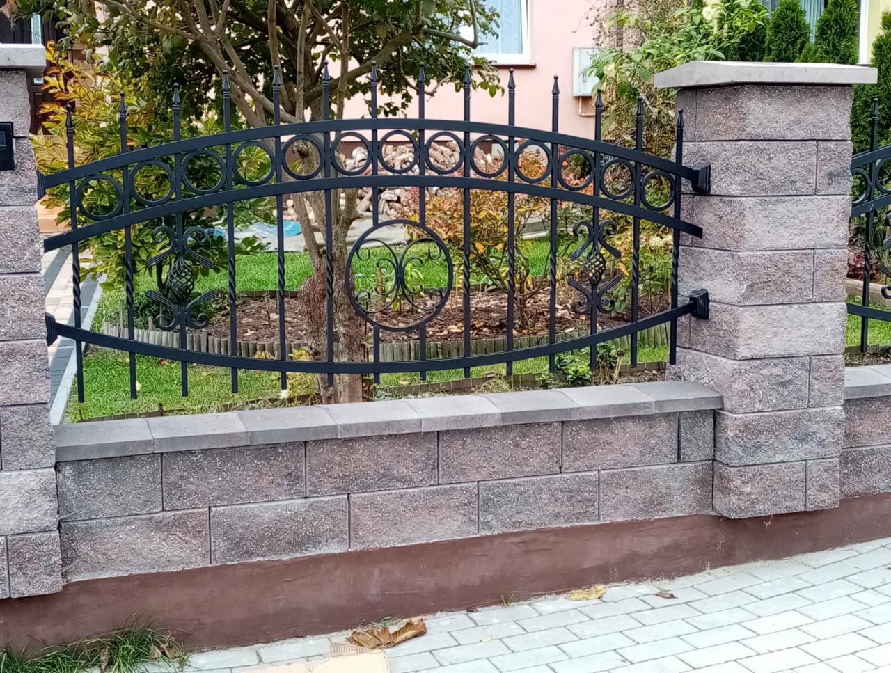 Fragment ogrodzenia z czarną metalową balustradą o dekoracyjnych elementach, osadzoną na murku z szarej kostki i słupkach z tego samego materiału, widok z zewnątrz na tle zieleni ogrodu.