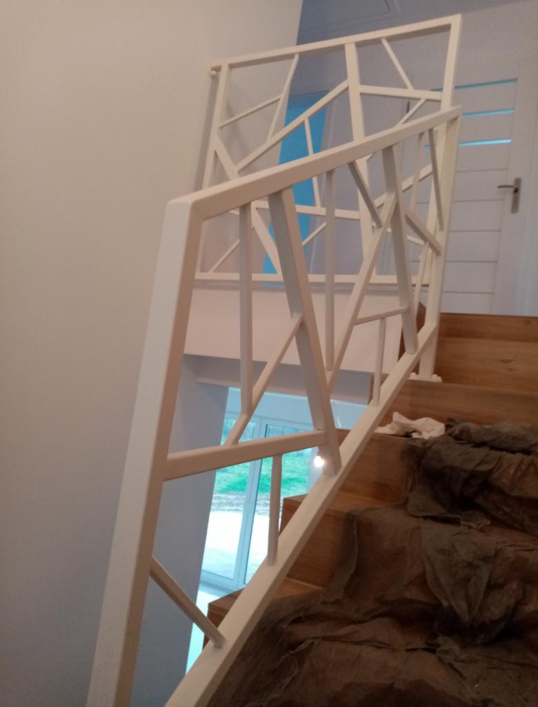 Biała, geometryczna balustrada schodowa z metalu, widok z góry na drewniane schody zabezpieczone folią malarską, w tle okno.