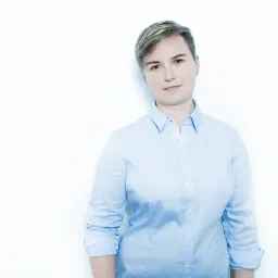 Portret kobiety w jasnoniebieskiej koszuli na białym tle, z krótkimi włosami i delikatnym uśmiechem.