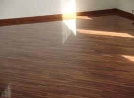 Błyszcząca, ciemnobrązowa podłoga z paneli, oświetlona promieniami słońca wpadającymi przez okno. Widoczne drewniane listwy przypodłogowe. Minimalistyczne wnętrze z białymi ścianami.