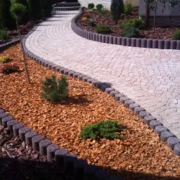 Zakrzywiona ścieżka z jasnej kostki brukowej, obrzeża z ciemnych betonowych palisad, rabata wysypana korą z iglakami, widok z góry.