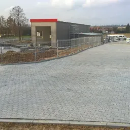 Ułożona szara nawierzchnia z kostki betonowej przy nowo powstającym budynku z czerwoną obudową, widoczny fragment terenu przygotowywany pod dalsze prace, ogrodzenie tymczasowe oraz zaparkowany...