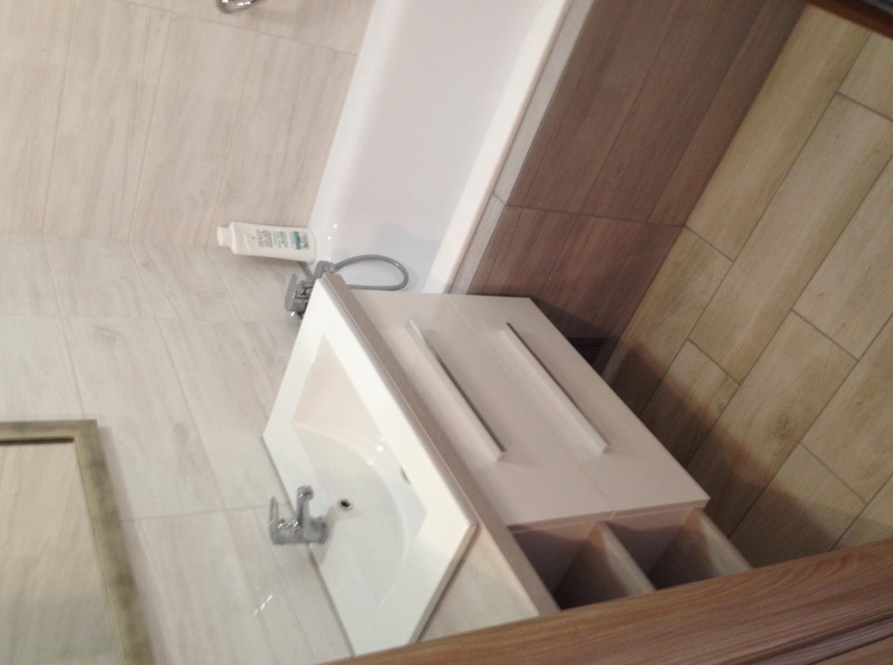 Nowoczesna biała umywalka z szafką w łazience wykończonej płytkami imitującymi drewno, widoczna bateria i fragment wanny.