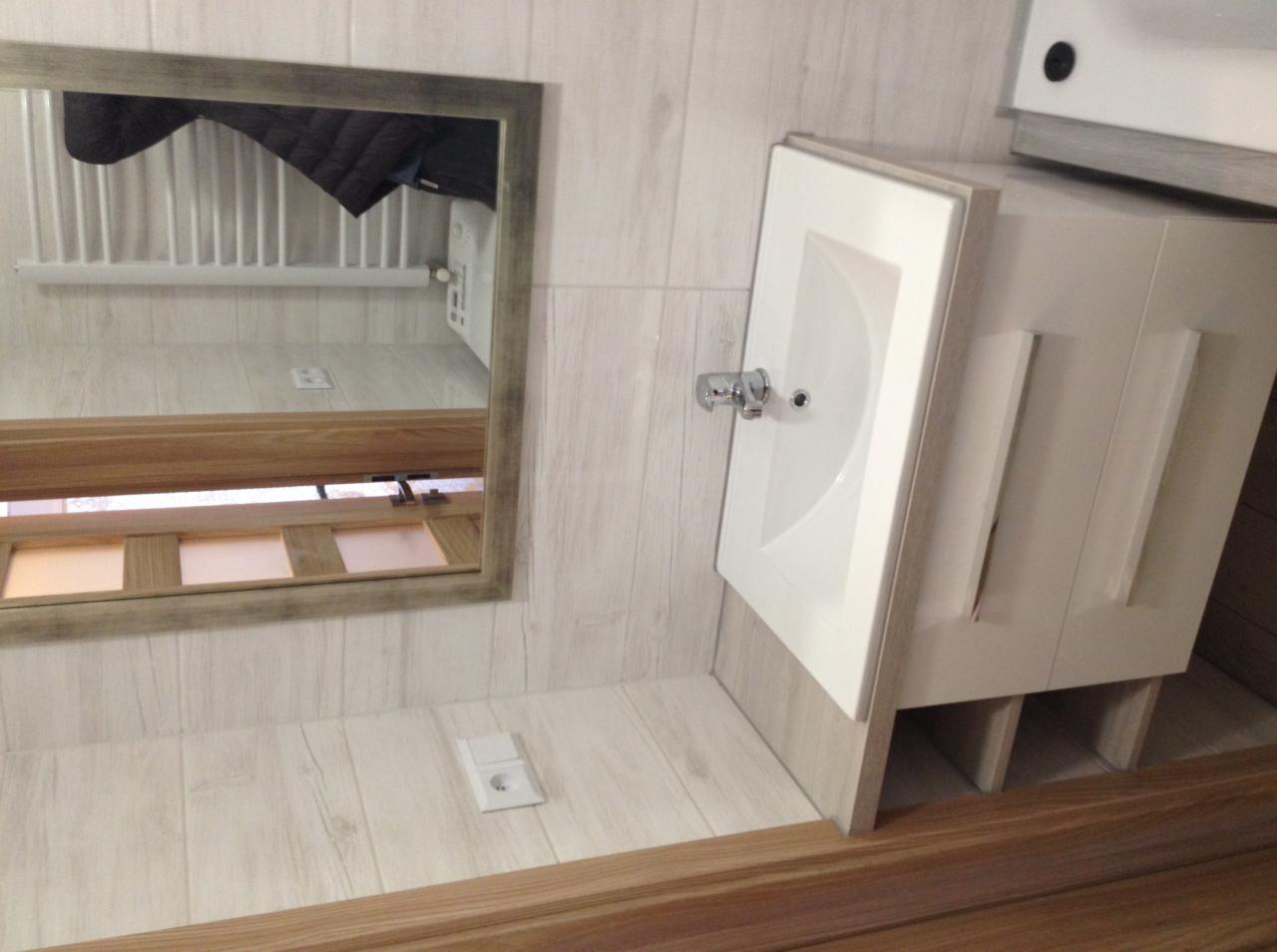 Biała umywalka z szafką w łazience z drewnopodobnymi płytkami, widoczne gniazdko elektryczne i lustro z odbiciem grzejnika.