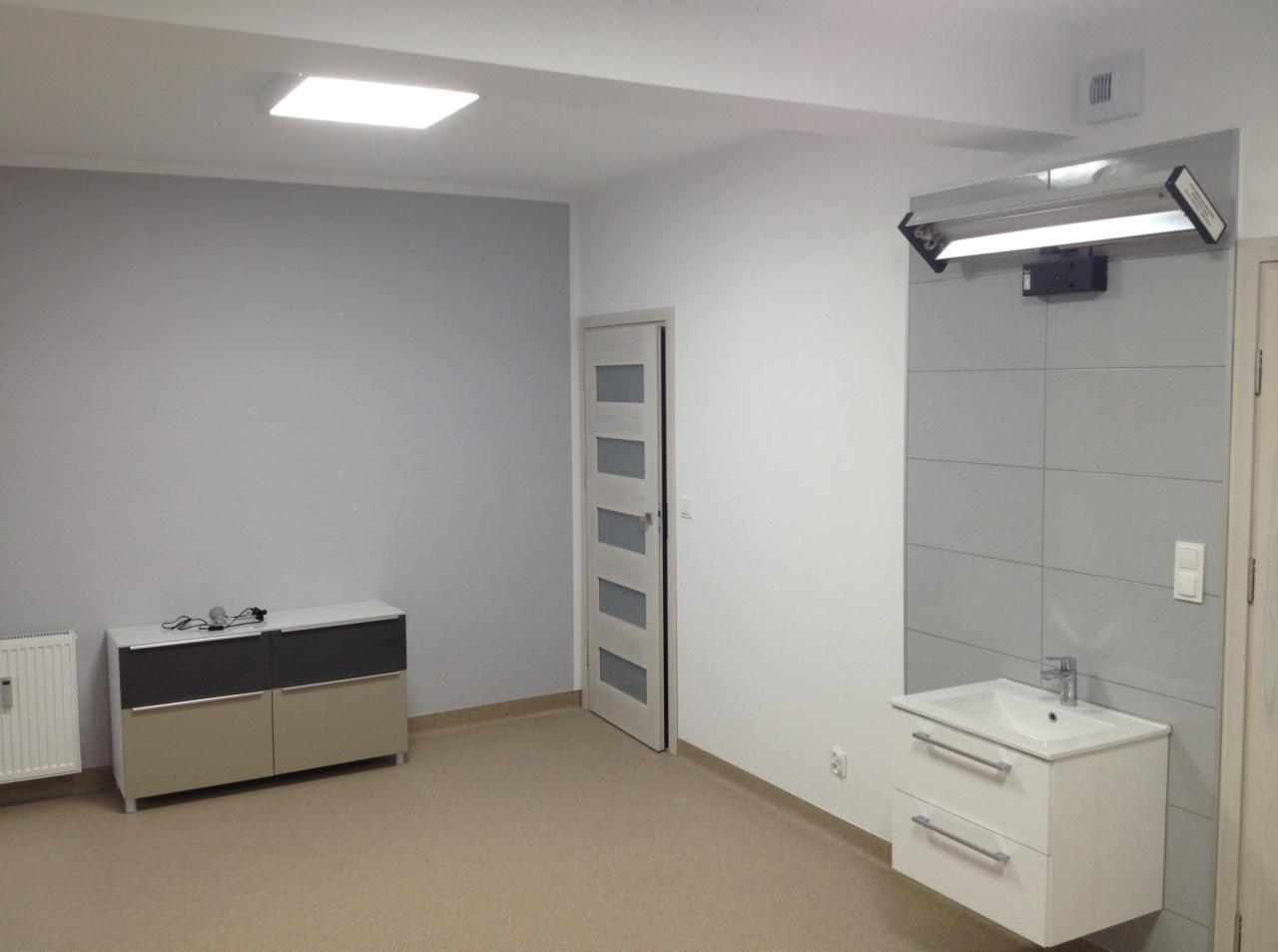 Szare płytki na ścianie z zamontowaną umywalką z szafką w nowym, jasnym pomieszczeniu z beżową podłogą i szarą ścianą, widoczny grzejnik, drzwi z przeszkleniami i lampa sufitowa.