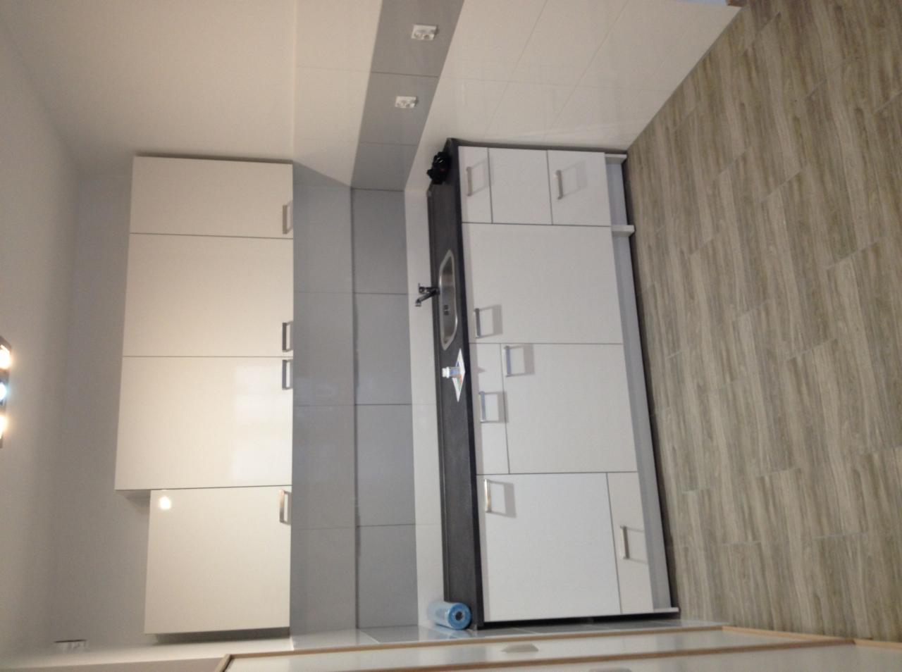 Nowoczesna łazienka z białymi szafkami i drewnopodobną podłogą, widok na umywalkę z baterią i rolkę niebieskiej folii zabezpieczającej na blacie.