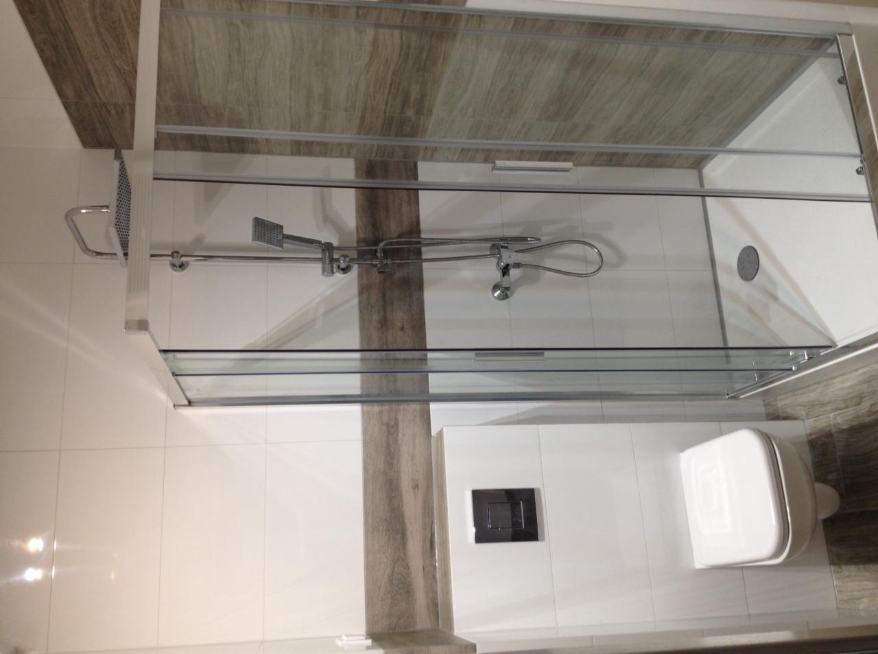 Nowoczesna kabina prysznicowa z przesuwnymi drzwiami szklanymi, zamontowana armatura prysznicowa z deszczownicą, widoczna podtynkowa spłuczka WC i wisząca toaleta w wykończonej łazience.