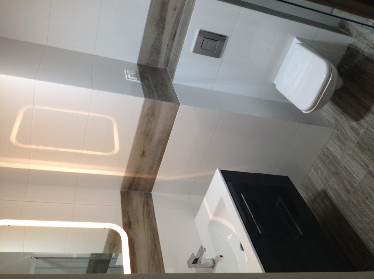 Nowoczesna łazienka z białymi płytkami, drewnopodobnymi akcentami, podwieszaną toaletą i szafką z umywalką. Ledowe oświetlenie lustra.