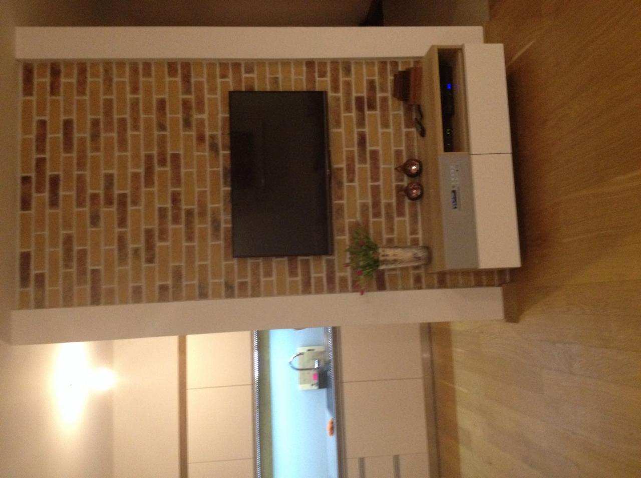 Telewizor zawieszony na ścianie wyłożonej cegiełkami klinkierowymi, obok szafka RTV z dekoracjami i kwiatami w wazonie, widoczny fragment blatu i frontów szafek kuchennych w tle.