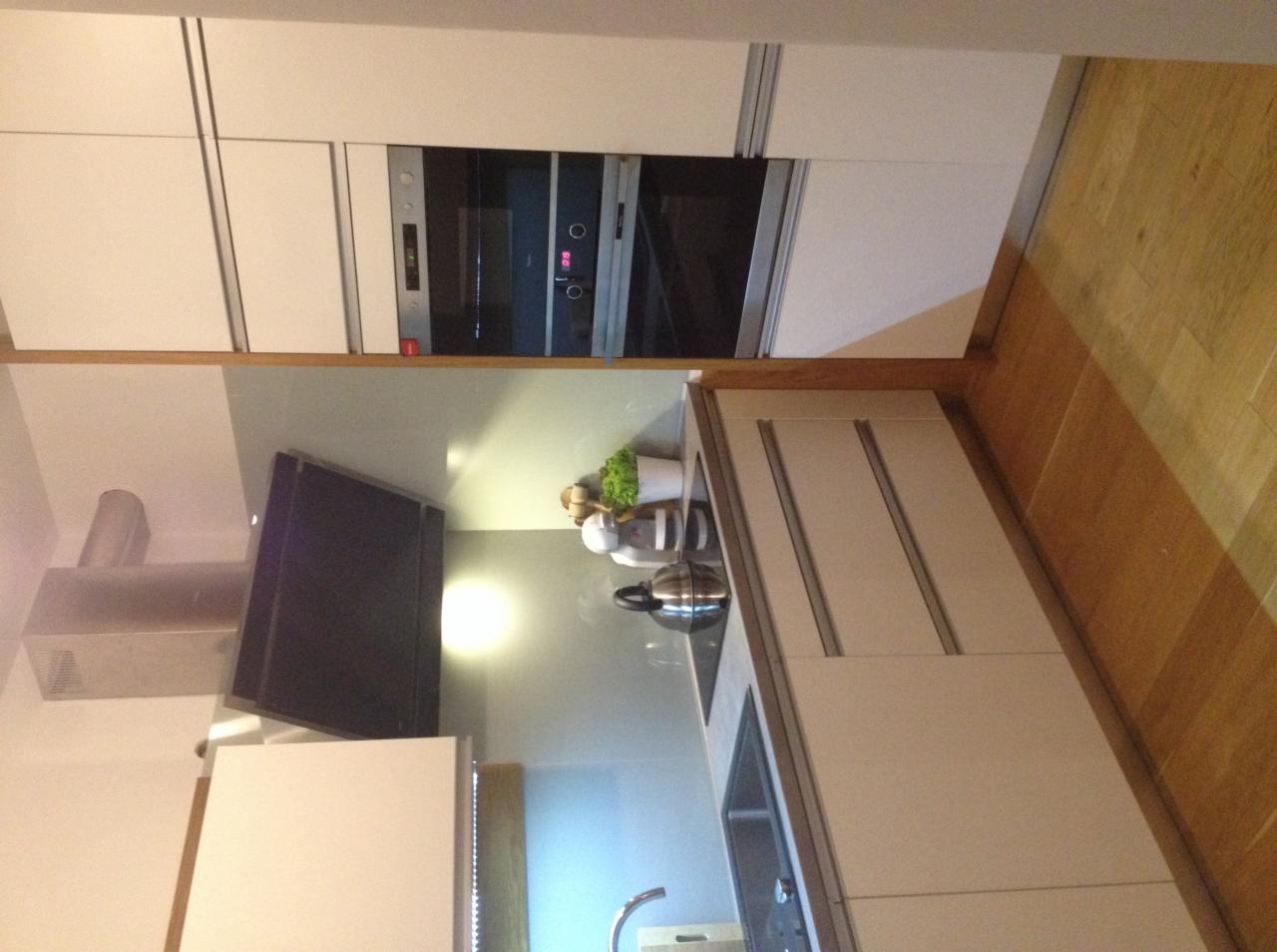 Nowoczesna, jasna kuchnia z zabudową meblową, piekarnikiem w zabudowie, okapem i drewnianą podłogą. Na blacie widoczny czajnik i dekoracje.